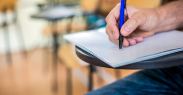A man is writing/signing on a paper. Focused on a hand with pen. undergraduate student holding pencil and sitting on row chair doing final exam attending in examination room or classroom.university student.